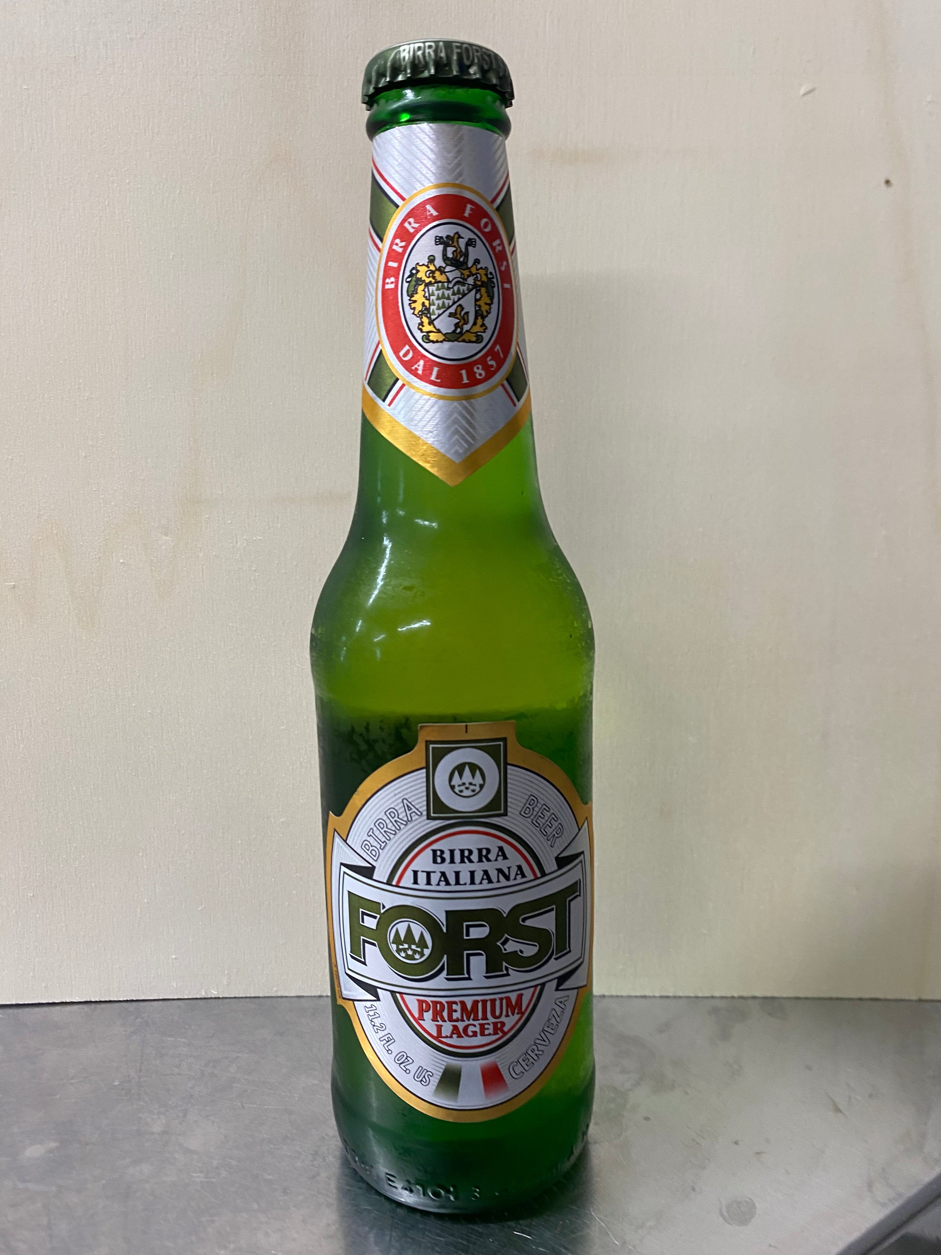 Green bottle of Forst Birra Italiana premium lager on a metallic surface with a light brown background