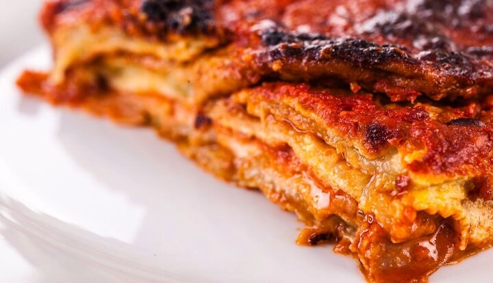 A close-up shot of a slice of lasagna on a white plate, showing layered pasta sheets, rich red tomato sauce, melted cheese, and a crispy dark brown baked crust.